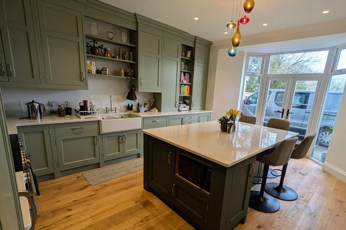 A sage green painted kitchen with an island, a bay window and coloured glass pendants