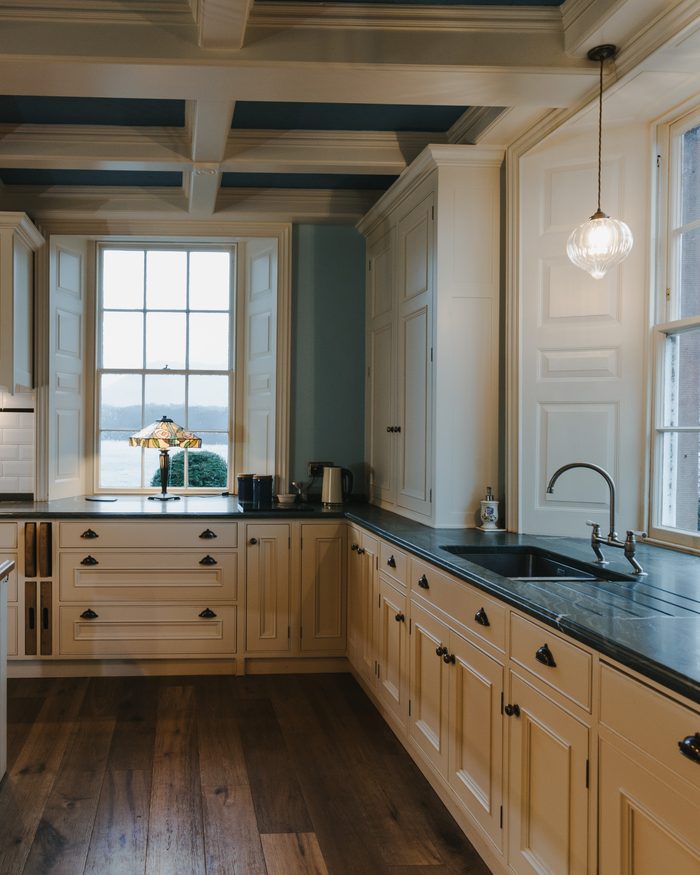 A cream painted kitchen with a coffered ceiling, panelled window shutters and dark stone worktops