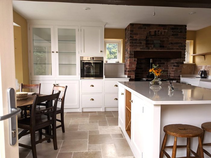 A white painted in-frame kitchen with a glazed dresser, an exposed brick inglenook and an island with a breakfast bar