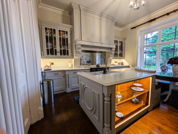 A pale grey painted country house kitchen with glazed wall cabinets, a carved mantel over the range and an island with a lit oak display end
